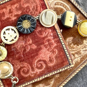 May include: A vintage button bracelet with a variety of buttons, including a black button with gold accents, a white button with a leaf design, a white button with a textured surface, a black button with two holes, and a yellow button with a swirl design.