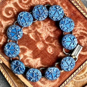 May include: A vintage bracelet featuring twelve round, blue buttons with a detailed design, connected by silver-toned rings. The buttons have a textured appearance and a decorative pattern. The bracelet is secured with a silver clasp.