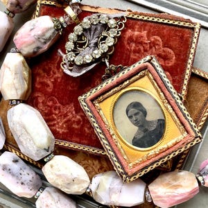 Puede incluir: Un relicario vintage con un pequeño retrato de una mujer en su interior, rodeado de un marco dorado y una caja de terciopelo rosa. El relicario cuelga de una cadena delicada con un dije de corazón de pedrería. También se puede ver un collar de cuentas rosas y blancas en la imagen.