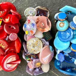 May include: A collection of vintage buttons in three columns. The first column is primarily red, the second is a mix of pinks, purples, and golds, and the third is various shades of blue. The buttons vary in size, shape, and material.