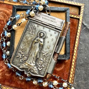 Könnte beinhalten: Vintage silberner religiöser Anhänger mit detailliertem Bild eines Heiligen, floralen Akzenten und dem Text "SOUVENIR DE LOURDES". Der Anhänger ist an einer Halskette mit blauen und weißen Perlen befestigt. Präsentiert in einer samtgefütterten Box.