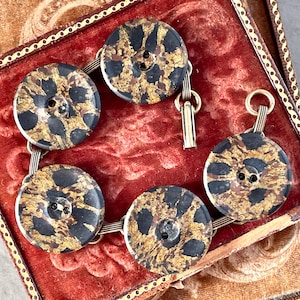 May include: A bracelet made of five round, black and gold buttons connected by a thin metal chain. The buttons have a speckled pattern and a small button in the center.