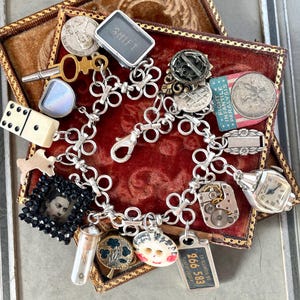 May include: A silver charm bracelet with a variety of charms, including a watch, a domino, a key, a coin, a small bottle, a button, a license plate, and a small picture frame with a black and white photo. The bracelet is on a red velvet background.
