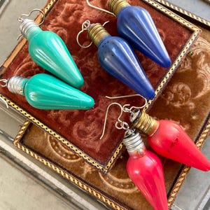 May include: Three pairs of dangle earrings made from light bulbs. One pair is teal green, one pair is blue, and one pair is red.