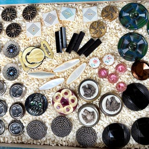 May include: A collection of vintage buttons in various shapes, sizes, and colors. The assortment includes metal, glass, and plastic buttons, some with intricate designs and embellishments. The buttons are arranged in a tray filled with a light-colored filler.