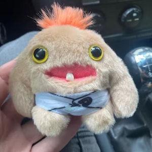 Peut inclure: Une peluche en forme de créature avec un corps beige, des cheveux orange et de grands yeux jaunes. Elle a une bouche rouge avec une seule dent et une couche bleue et blanche. La peluche est douce et duveteuse.