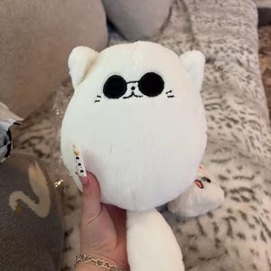 May include: A white plush toy cat with black eyes, whiskers, and small ears. The cat has a round body and long, dangling legs. The toy is made of soft, fluffy material.