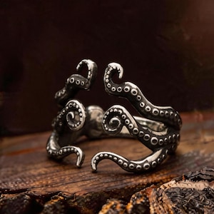 Puede incluir: Un anillo plateado con un diseño de pulpo. Los tentáculos del anillo se envuelven alrededor del dedo, con ventosas detalladas visibles. El anillo está sobre una superficie de madera rústica, resaltando el diseño.