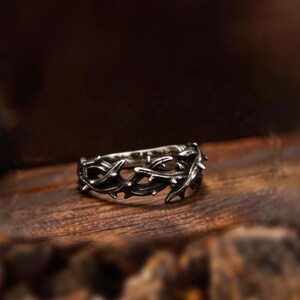 Könnte beinhalten: Ein silberfarbener Ring mit einem dornigen Rankenmuster. Der Ring ist vor einem dunklen, verschwommenen Hintergrund platziert, der die komplizierten Details der Metallarbeiten hervorhebt. Das Design des Rings weist spitze Dornen auf.
