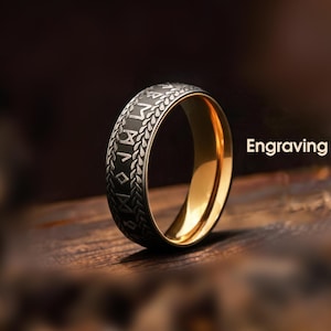 May include: A close-up of a ring with a gold interior and a dark, engraved exterior. The ring features a pattern of symbols and text, with the word "Engraving" visible in the background. The ring is set against a blurred, dark background.