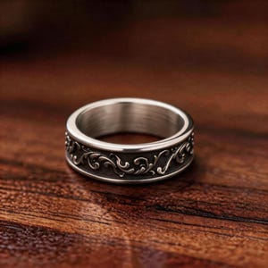 May include: A silver-toned metal ring featuring a wide band with a recessed centre. The band displays a raised, intricate scrollwork pattern in a metallic finish against a darkened, oxidised background, creating a high-contrast visual effect.