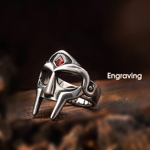 May include: A silver-colored ring with a mask design, featuring a red gemstone. The ring has a detailed, engraved appearance and the word "Engraving" is displayed in white text.