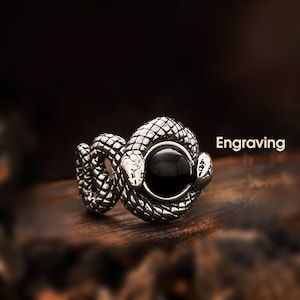 May include: A silver-toned ring featuring a coiled snake design with a large, round black stone at its center. The snake's head is visible on either side of the stone. The word "Engraving" is printed in white.