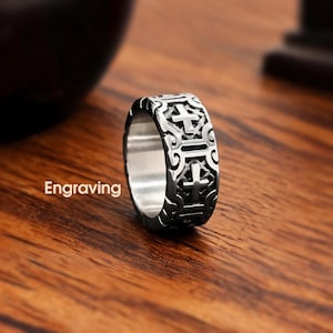 Peut inclure: Une bague en métal argenté présentant un motif répétitif de croix gravées et de volutes complexes. Le mot Engraving est imprimé en blanc à gauche de la bague, qui possède une surface intérieure de bande lisse et polie.