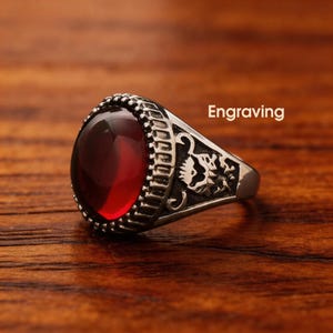 May include: A silver-toned ring featuring a large, smooth, oval-shaped deep red gemstone set in a bezel with a textured edge. The side of the ring band displays an intricate engraved design. The word Engraving is visible in the image.