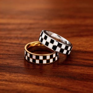 May include: Two rings with a checkerboard pattern. One ring is gold-toned, and the other is silver-toned. Both rings have a band of alternating black and white squares around the circumference. The rings are stacked on a dark wood surface.