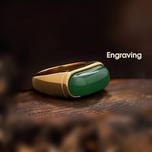 May include: A gold ring with a rectangular, emerald-green stone. The ring has a polished gold band and a smooth, rounded top. The word "Engraving" is displayed in white text.