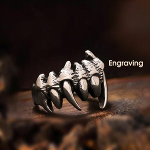 May include: A silver-colored ring with a detailed design of sharp teeth. The ring has a dark background and the word "Engraving" is displayed in white text. The ring is a unique piece of jewelry.
