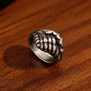 May include: A silver-toned metal ring featuring a detailed design of two hands clasped together in a handshake. The surface of the ring has a darkened, oxidised finish that highlights the texture and contours of the fingers and knuckles.