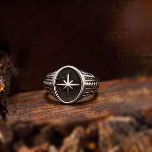 May include: A silver ring with an oval face featuring a compass rose design. The ring has a dark, oxidised finish and detailed side accents. The ring is displayed on a wooden surface.