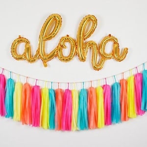 May include: Gold foil "aloha" balloon letters above a colorful tassel garland. The garland features bright pink, turquoise, yellow, and orange tassels, strung on a thin string, creating a festive and vibrant party decoration.