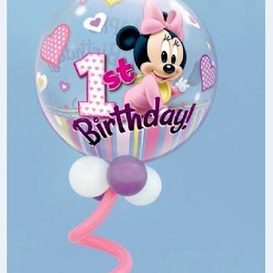 May include: A clear bubble balloon featuring Minnie Mouse in a pink outfit and bow. The balloon has a "1st Birthday!" message and is decorated with hearts. The balloon is supported by a pink curly stem and white and purple balloons.