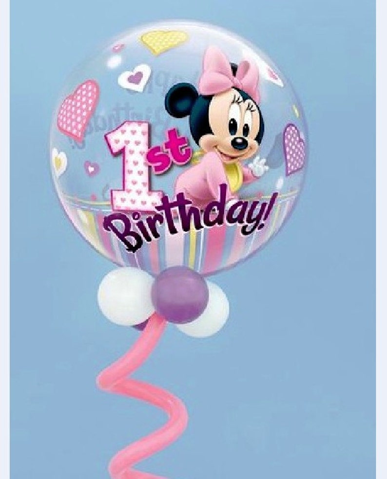 May include: A clear bubble balloon featuring Minnie Mouse in a pink outfit and bow. The balloon has a "1st Birthday!" message and is decorated with hearts. The balloon is supported by a pink curly stem and white and purple balloons.