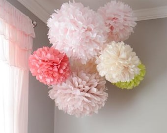 May include: A cluster of paper pom-poms in various shades of pink, coral, and cream, with a touch of green. These decorative, fluffy spheres are arranged together, creating a soft, whimsical aesthetic. The pom-poms are likely used for party or room decor.