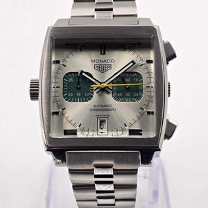 Op de afbeelding: Zilverkleurig Monaco Heuer automatisch chronograaf horloge met een vierkante wijzerplaat en een roestvrijstalen band. De wijzerplaat heeft groene en gele accenten, met de datum onderaan. Swiss Made.