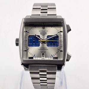 Könnte beinhalten: Silberne Monaco Heuer Automatik-Chronographenuhr mit quadratischem Zifferblatt und Edelstahlarmband. Das Zifferblatt hat blaue und silberne Details, mit Datumsanzeige. Swiss Made.