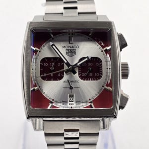 May include: A stainless steel TAG Heuer Monaco automatic watch. The square watch face has a silver dial with red accents and black hands. The watch band is also stainless steel.