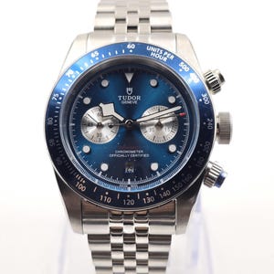 Può includere: Un orologio cronografo in acciaio inossidabile color argento con quadrante blu soleil, caratterizzato dal logo Tudor Geneve, due sotto-quadranti argentati, una finestrella della data a ore sei e una lunetta tachimetrica blu su bracciale in metallo.