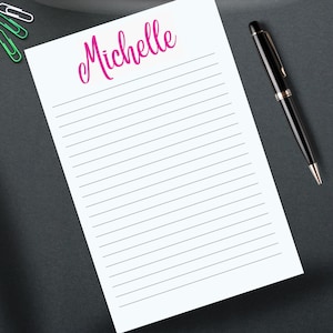 Può includere: Un blocco note bianco a righe con il nome "Michelle" in corsivo rosa in alto. Una penna nera con dettagli dorati poggia sul lato destro del blocco note. Graffette verdi nell'angolo in alto a sinistra. Il blocco note è su una superficie grigio scuro.