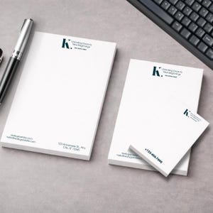 Puede incluir: Blocs de notas blancos de varios tamaños con el texto "Unlocking Doors to New Beginnings" y un logotipo "K" turquesa. Los blocs de notas también incluyen información de contacto. Un bolígrafo y un teclado son visibles en el fondo.