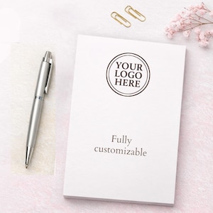 Puede incluir: Un bolígrafo plateado junto a un bloc de notas rectangular blanco. El bloc presenta un gráfico circular con el texto YOUR LOGO HERE y la frase Fully customizable impresa debajo. Dos clips dorados están colocados sobre el bloc de notas.