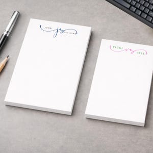 Può includere: Due blocchi per appunti bianchi con testo personalizzato. Un blocco per appunti ha il nome "John Sullivan" in corsivo blu navy. L'altro blocco per appunti ha il nome "Vicki Sell" in corsivo rosa e verde. Sono visibili una penna e una matita.
