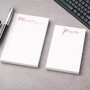 Peut inclure: Deux blocs-notes blancs personnalisés. L'un porte le texte "mc MELISSA CHRISTINE" en rose, l'autre "jc JORDAN CORUM" en noir. Un stylo argenté et noir et un clavier noir sont également visibles.