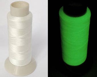 May include: A spool of white thread on a light beige base. The thread is tightly wound around a cylindrical core. In a second image, the thread glows bright green in the dark, highlighting its luminous quality. Suitable for sewing and crafting.