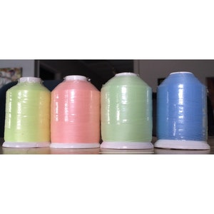 May include: Four spools of thread in pastel colors: yellow, pink, green, and blue. Each spool is wrapped in clear plastic and sits on a white base. The thread is tightly wound, suggesting a high-quality product for sewing or embroidery.