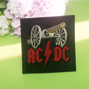 Könnte beinhalten: Schwarzer quadratischer Aufnäher mit einem gestickten Kanonenmotiv und dem Text "AC/DC" in Rot. Ein roter Blitz befindet sich in der Mitte. Der Aufnäher ist für Kleidung oder Accessoires bestimmt.