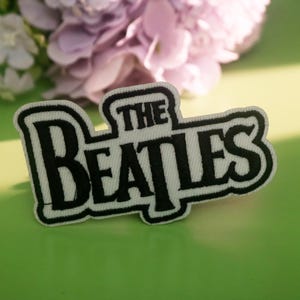 May include: An embroidered patch with the words "THE BEATLES" in black on a white background. The patch is edged in white and sits on a green surface, with a floral arrangement of purple flowers in the background.