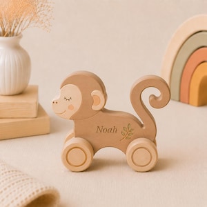 May include: A light brown wooden monkey toy on wheels featuring a curved tail and a smiling face with closed eyes. The side of the monkey is engraved with the name Noah and a small leaf sprig design. The toy has a smooth finish and four wheels.