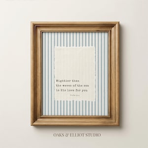 May include: Framed print with a light brown wooden frame. The print features a light blue and white striped background, a textured off-white paper with the text "Mightier than the waves of the sea is His love for you" and the text "Psalm 91:4".