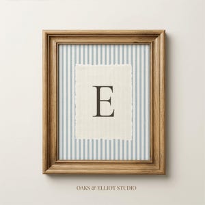 May include: A framed wall art piece featuring a light blue and white vertical striped background. Centered is a rectangular piece of off-white fabric with deckled edges, displaying the dark brown capital letter E. The text OAKS & ELLIOT STUDIO appears below.