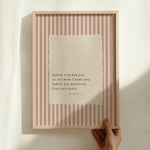 May include: A framed print with a light wood frame. The background has vertical pink and white stripes. A textured, off-white rectangle in the center contains the text "Before I formed you...Jeremiah 1:5."