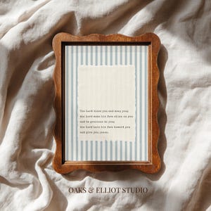 May include: A framed print with a wooden, wavy-edged frame. The print features a light blue and white striped background with a textured, off-white rectangle in the center. Text reads, "The Lord bless you and keep you...and give you peace."