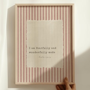 Può includere: Stampa artistica incorniciata con una cornice in legno chiaro. Lo sfondo presenta un motivo a righe rosa e bianche. Una carta strutturata, bianco sporco, con il testo "I am fearfully and wonderfully made" è centrata. Salmo 139:14 è stampato sotto.