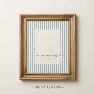 May include: A framed wall art piece featuring a light blue and white vertical striped background. Centered is a rectangular piece of off-white fabric with torn edges, displaying the black printed text: For this child I prayed 1 Samuel 1:27.