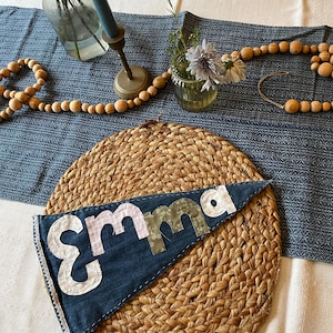 May include: A denim pennant with the name "Emma" in white, pink, and green letters, sits on a woven placemat. The pennant is trimmed with white lace. The scene includes a blue tablecloth, wooden beads, and floral arrangements.