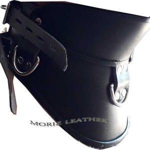 Op de afbeelding: Zwarte leren kraag met metalen D-ringen en verstelbare riemen. De kraag heeft een uitlopend ontwerp en is vastgezet met klinknagels. De tekst "MORIZ LEATHER" is zichtbaar aan de onderkant van de kraag.